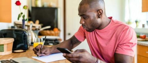 Man writing on a piece of paper while using a laptop and cell phone
