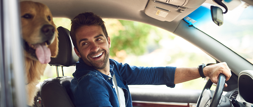 Man sitting in front seat of his car smiling at his dog in the backseat.