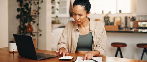 Woman reviewing receipts with a calculator and laptop at her kitchen table.